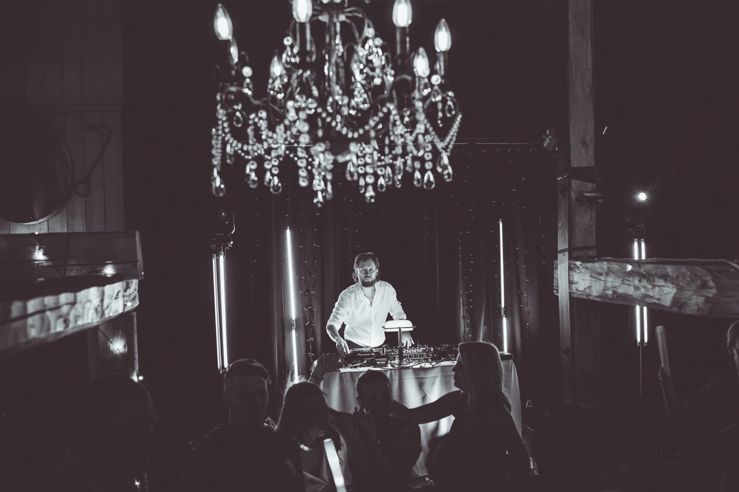 DJ i Trondheim ved miksepulten under en krystallysekrone i en tømmerlåve, med gjester i silhuett på dansegulvet foran