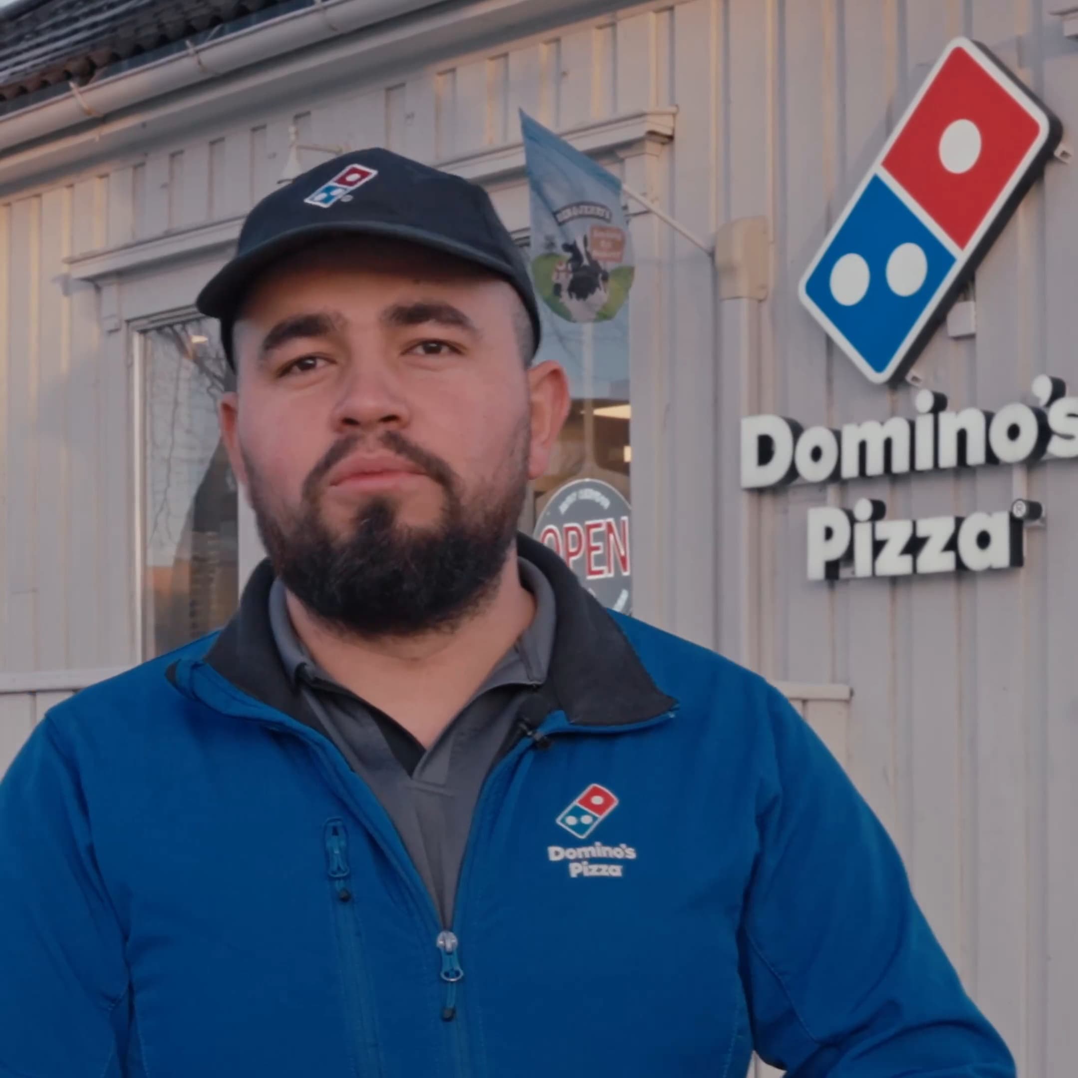 En ansatt hos Domino's snakker til kamera utenfor restauranten, med Domino's Pizza-skiltet og en OPEN-skilt i bakgrunnen