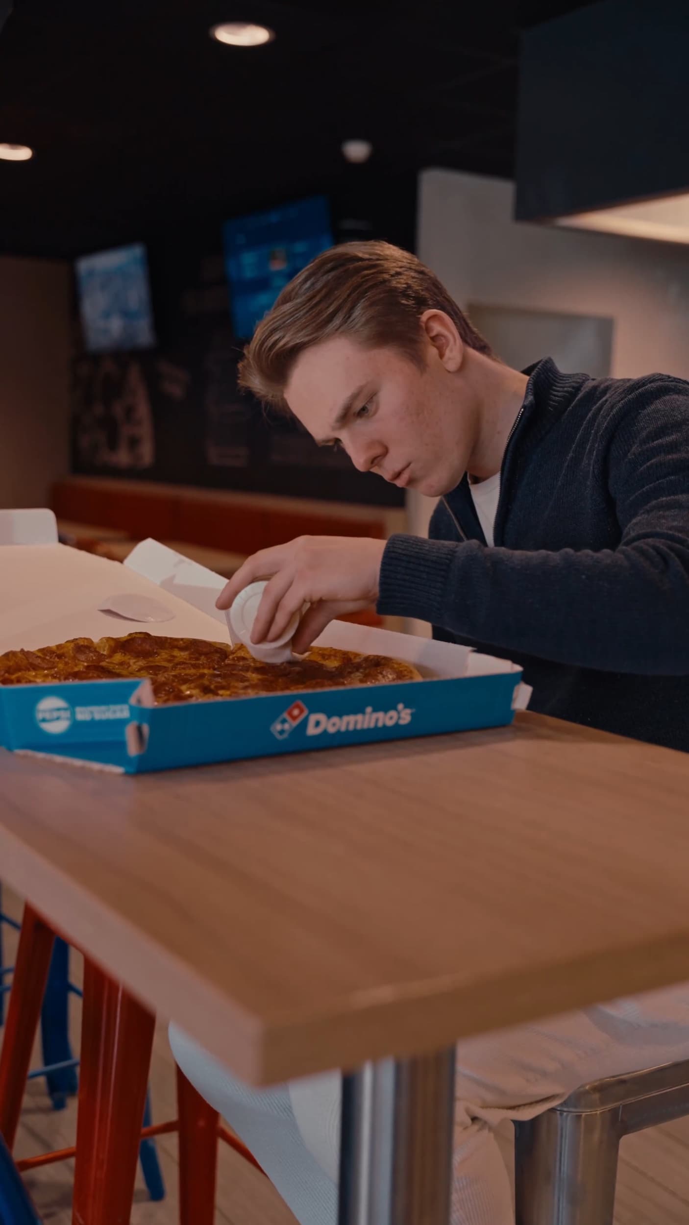 En gjest åpner en Domino's-pizzaeske og heller hvitløksdip over pizzaen ved et bord i restauranten