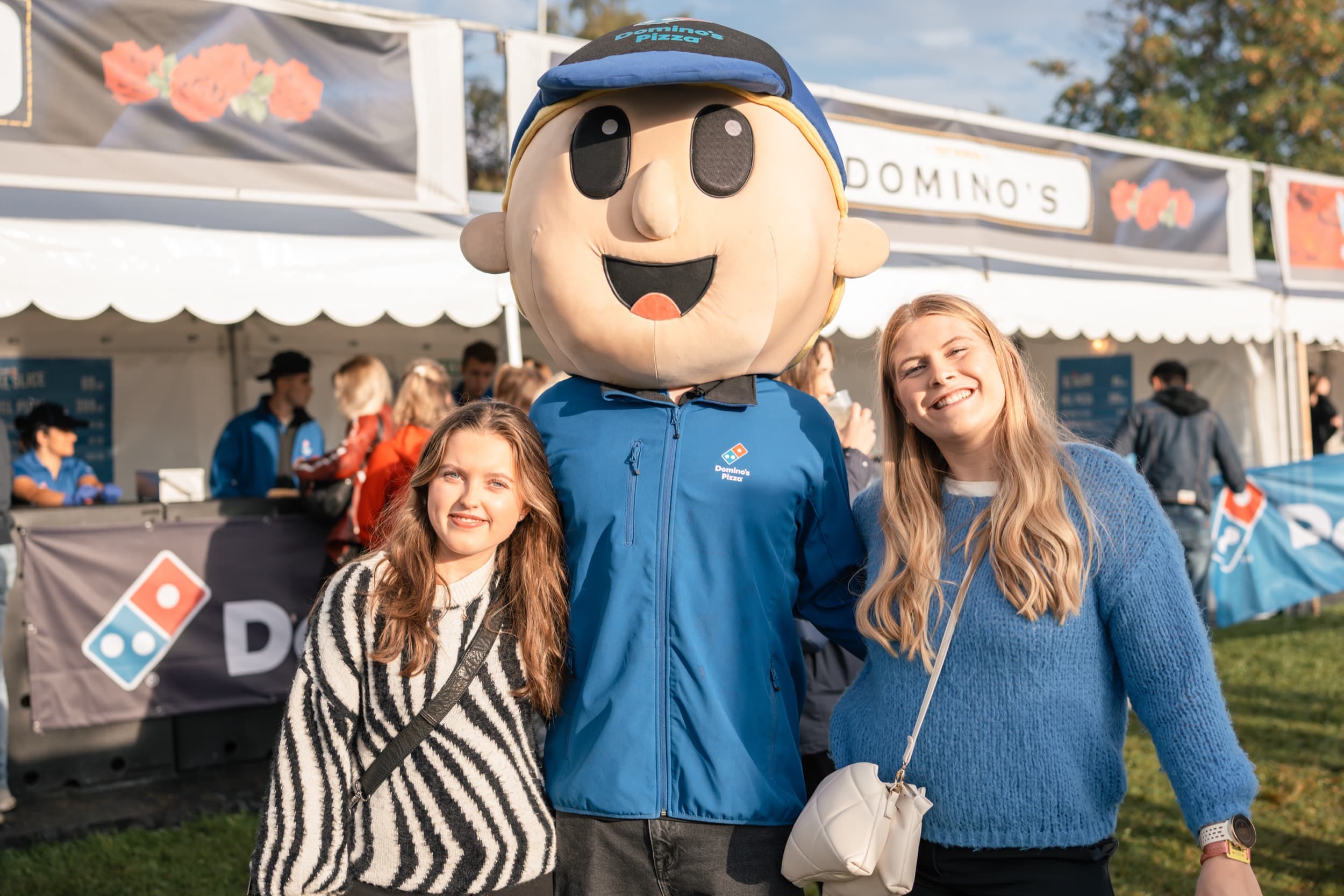 Domino's-maskoten poserer med to festivalgjester foran Domino's sin stand på Festningen, med Domino's Pizza-banner og flagg synlig.