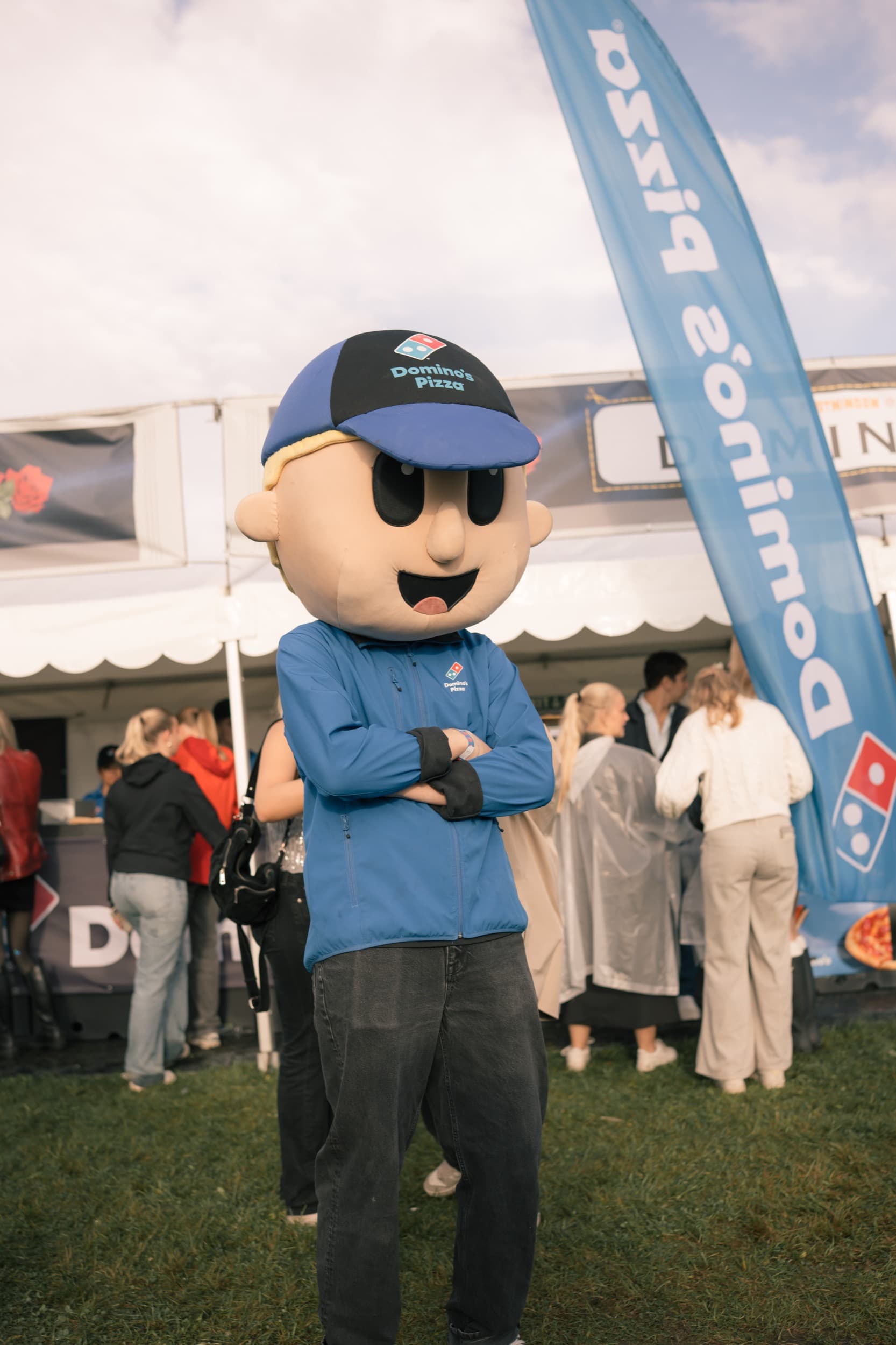 Domino's-maskoten står med armene i kors foran Domino's-standen på Festningen, med «Domino's Pizza» synlig på caps og jakke.