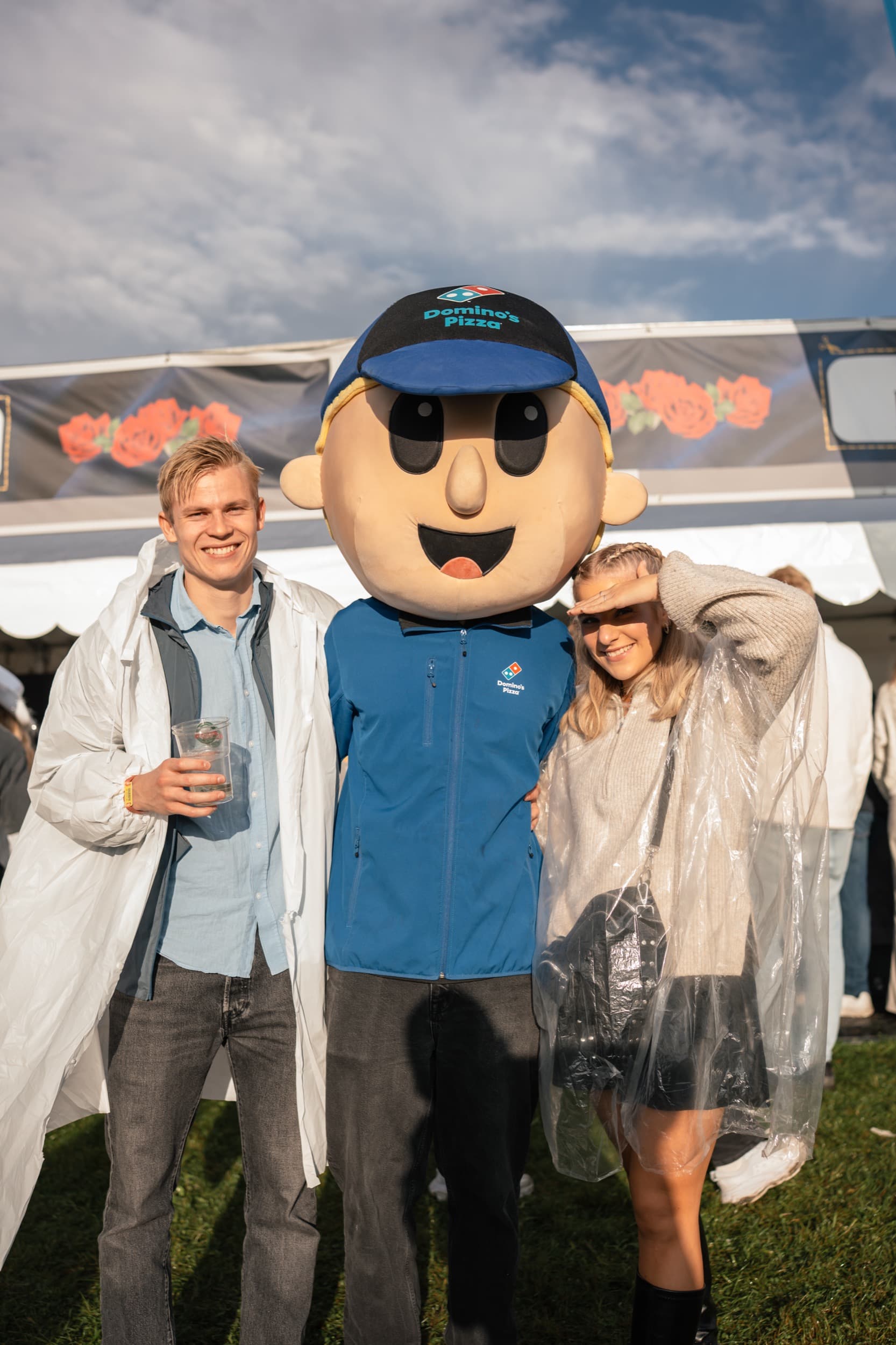 To festivalgjester i regnponcho poserer sammen med Domino's-maskoten foran standen i kveldslys.
