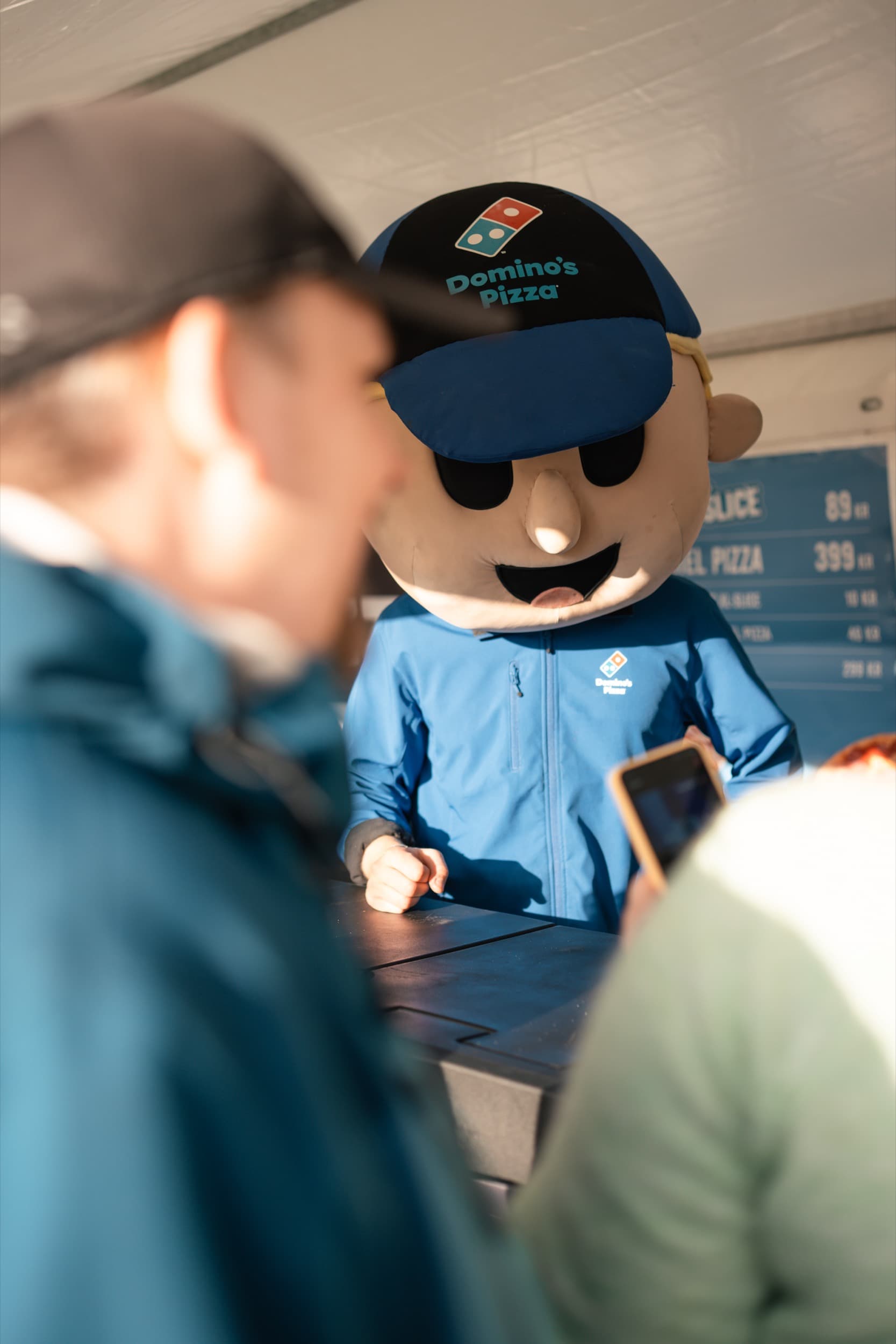 Domino's-maskoten ved disken i standteltet, med prisskilt for pizza synlig i bakgrunnen.