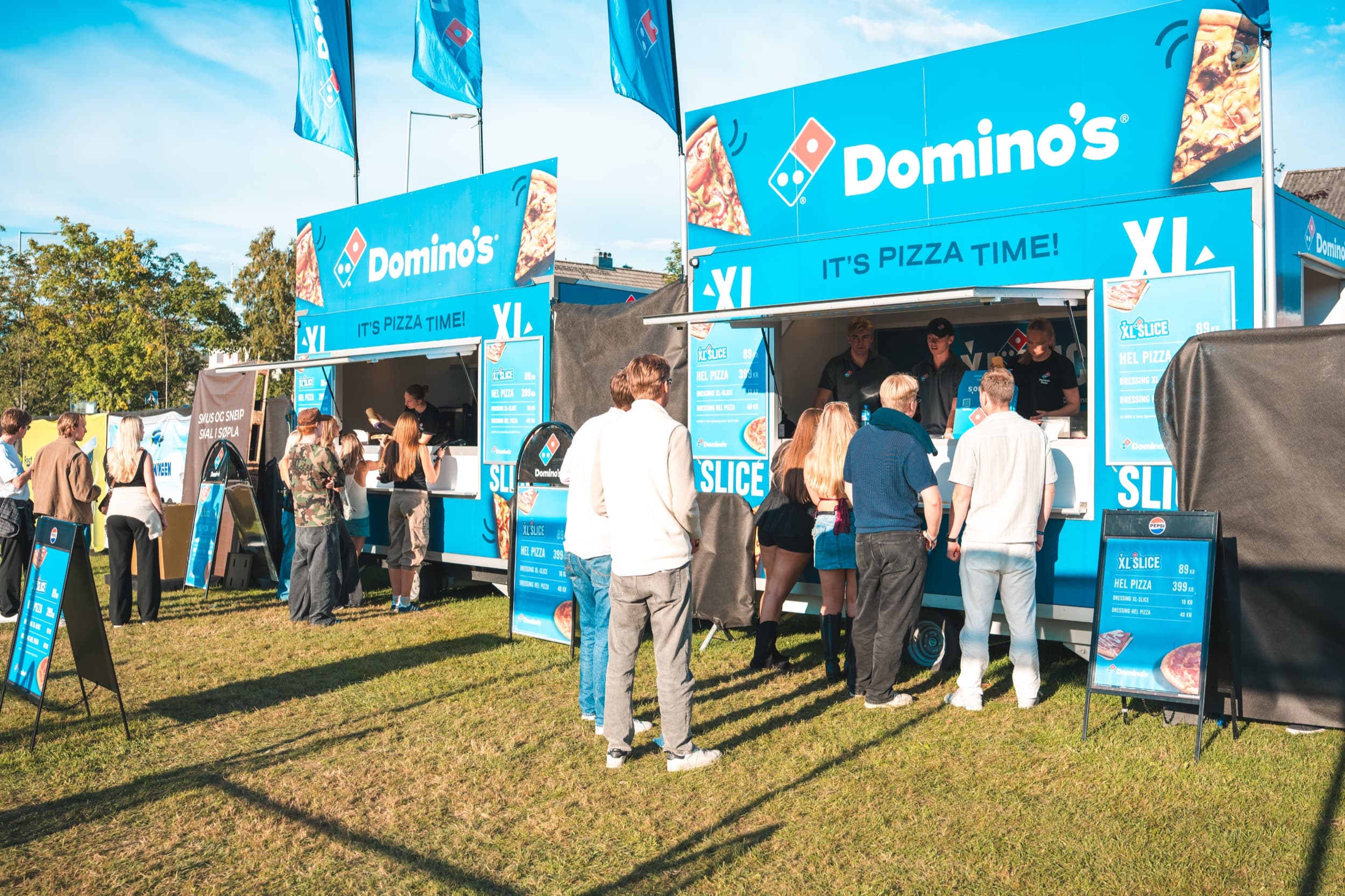 To Domino's-matvogner med flagg og «It's Pizza Time!»-branding på Festningen, med kø av gjester og crew synlig i lukene