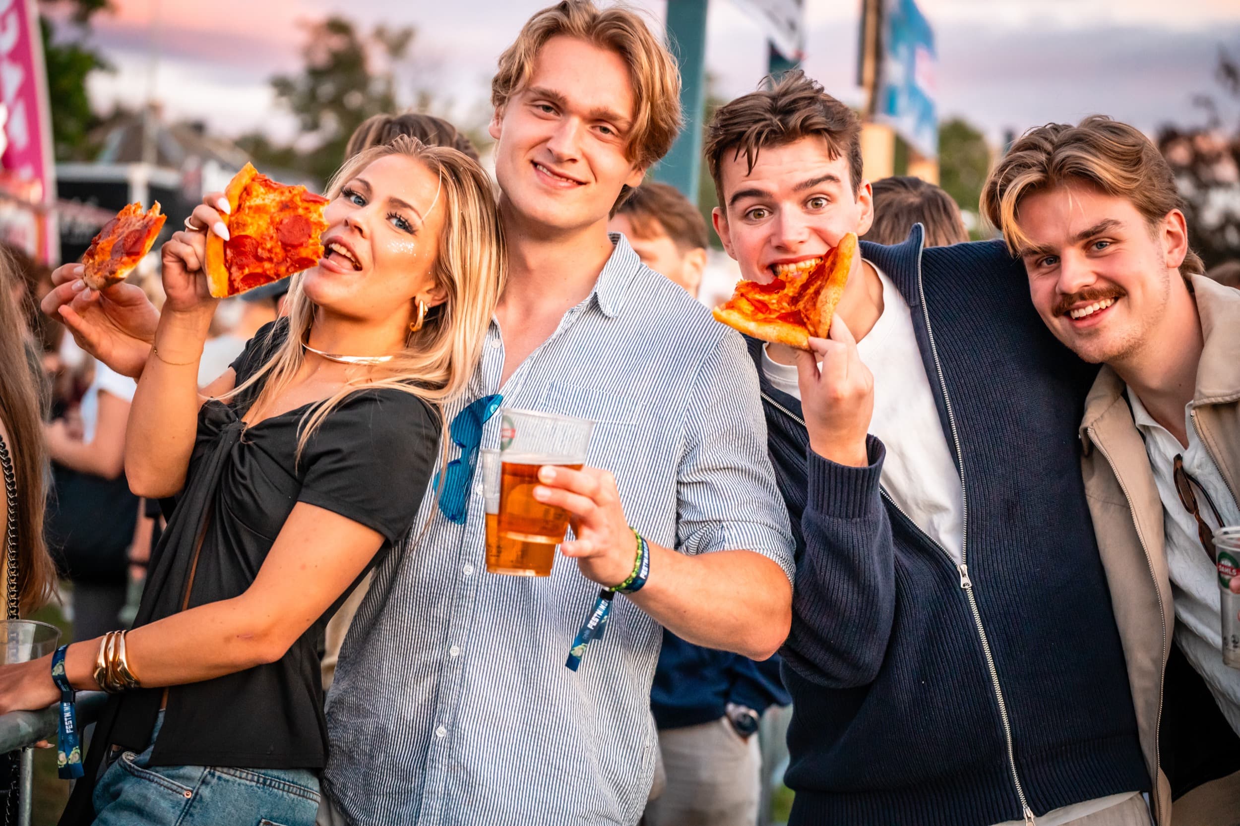 En vennegjeng spiser pizzaskiver og skåler med plastglass foran Domino's-flagg på Festningen
