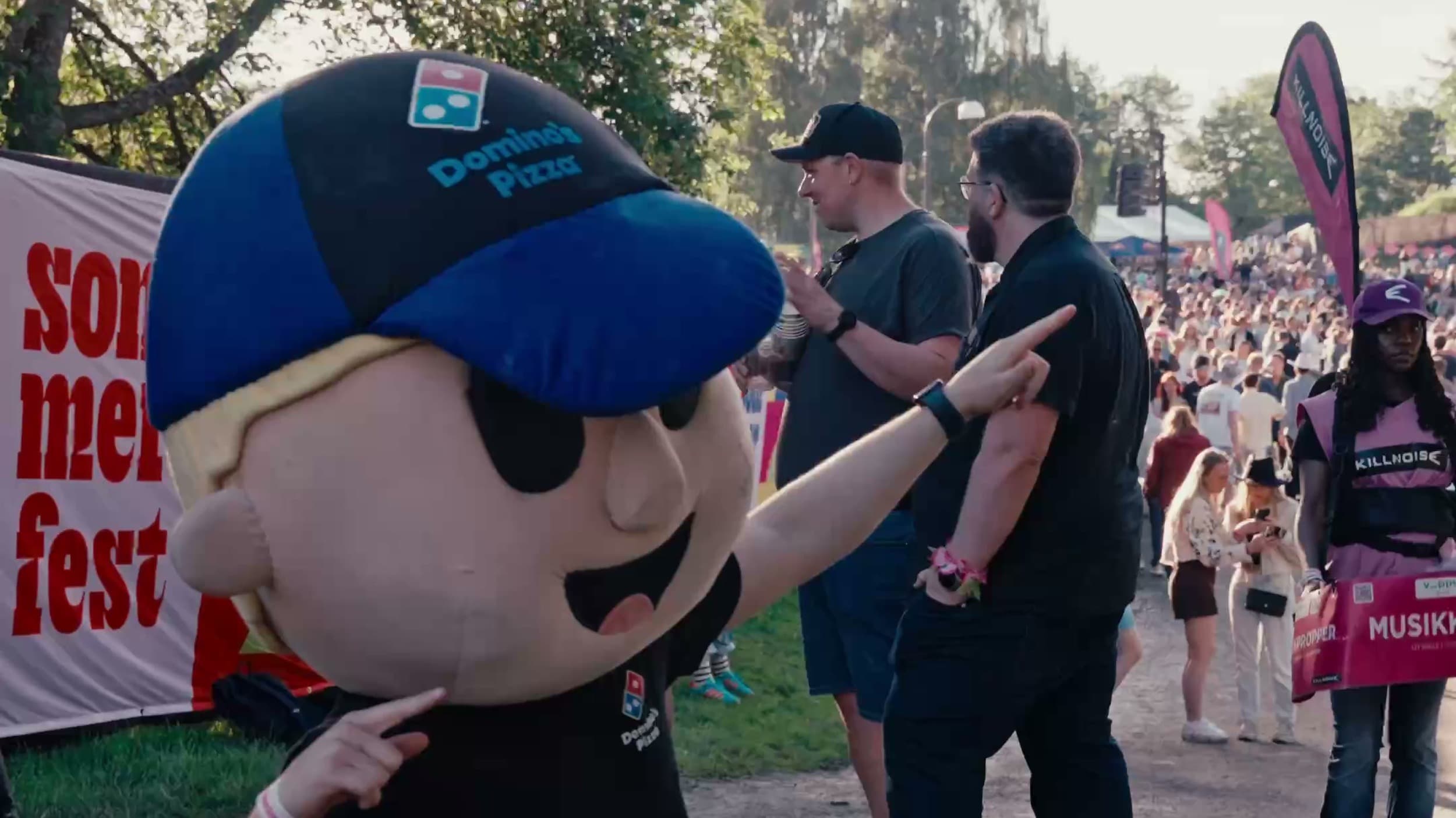 Domino's-maskoten kneler og peker mot kameraet foran «sommerfest»-skiltet, med festivalpublikum i bakgrunnen