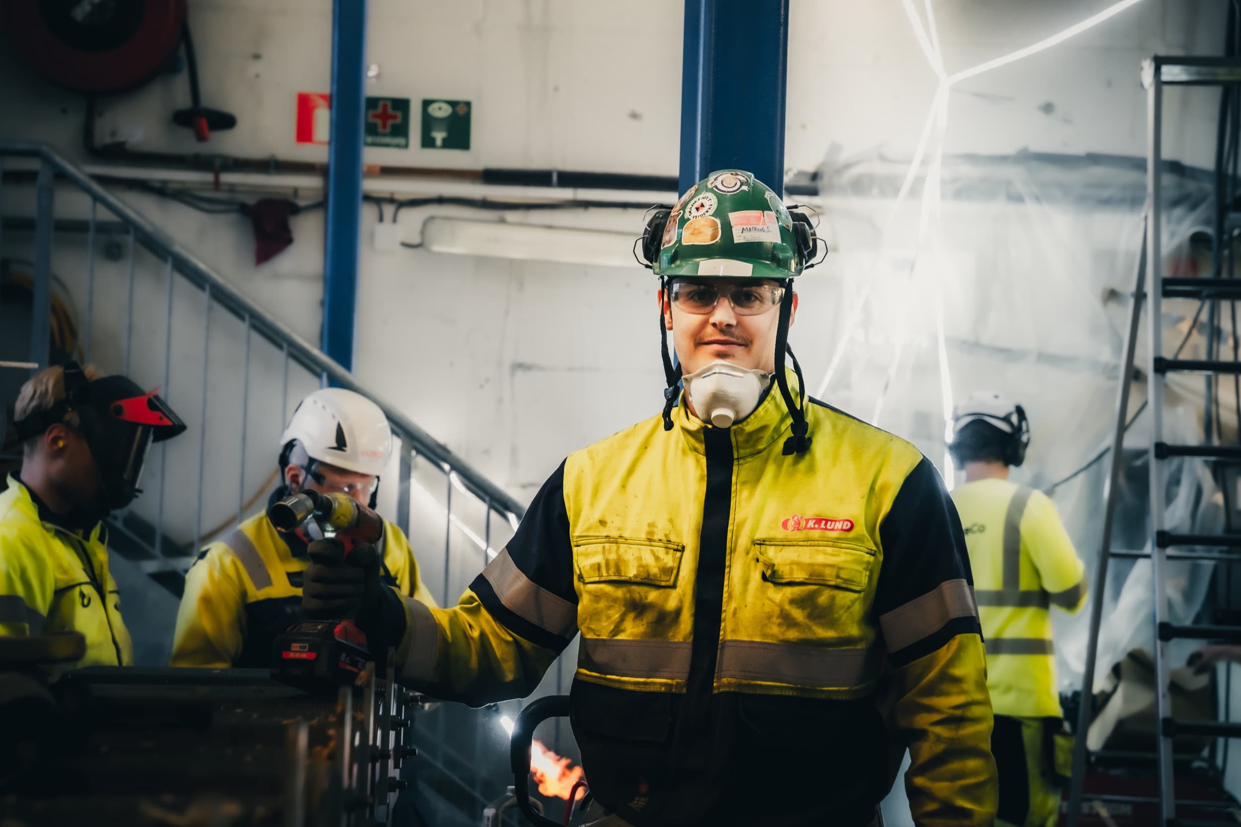 Ansatt i K. Lund med hjelm og vernebriller, med selskapets logo synlig på jakken, fotografert blant kolleger i arbeid