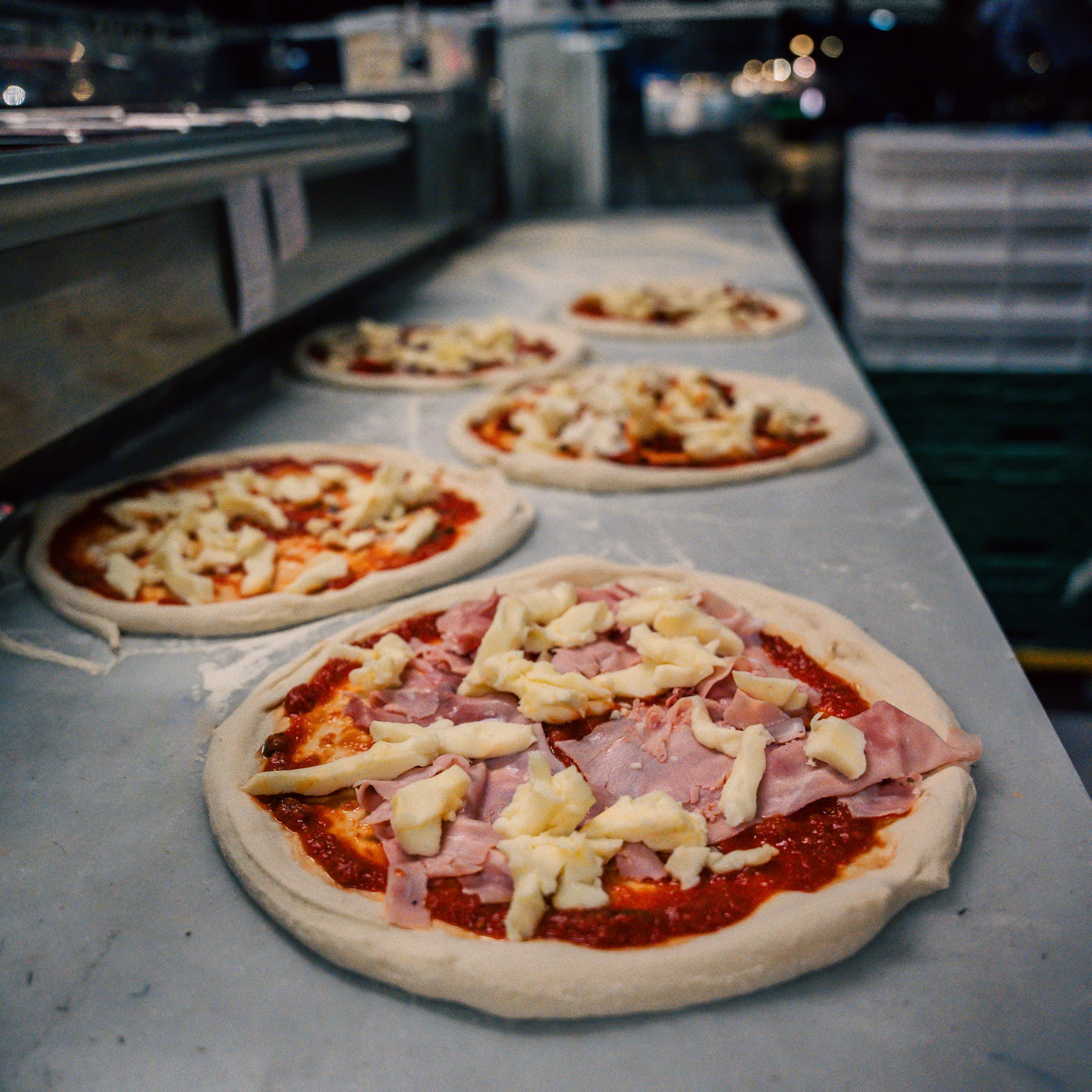 Ferdig kledde pizzaer klare for steking, med lys i bakgrunnen om kvelden