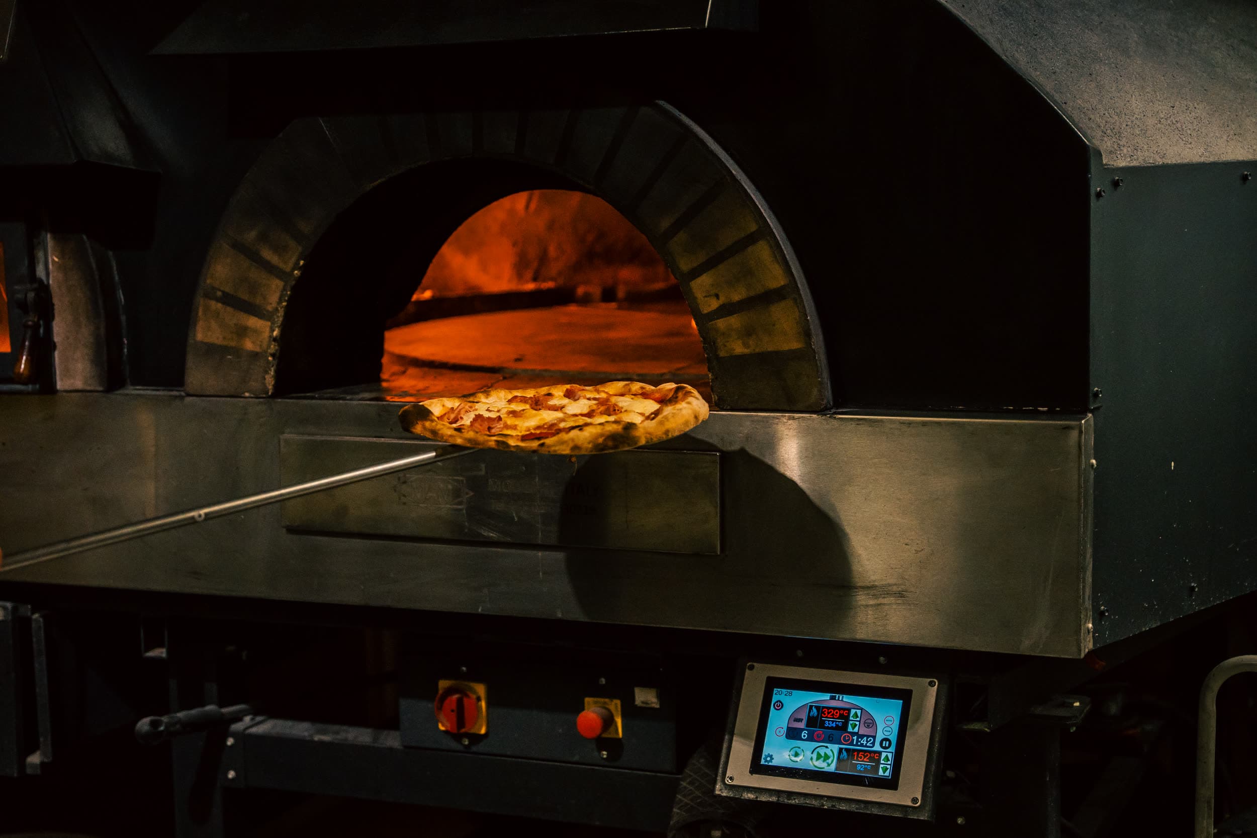 Pizza på ovnsspade settes inn i en vedfyrt pizzaovn med flammer synlig